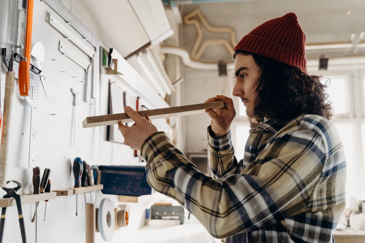 A young man in a joinery workshop holds up a length of timber to check it is straight, with hand tools hanging on the wall beside him.
