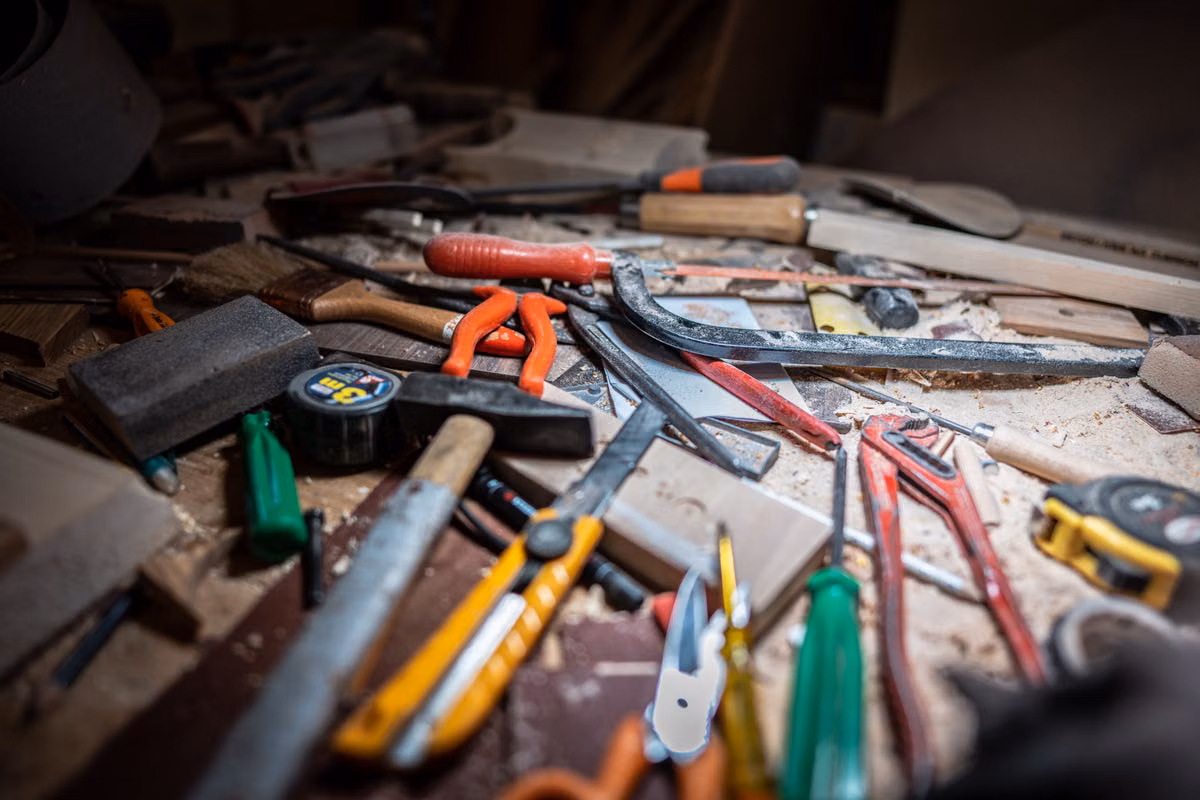 A close-up of hand tools, including saws, pliers, a hammer, screwdrivers, utility knives and tape measures, scattered across a dusty workshop bench.