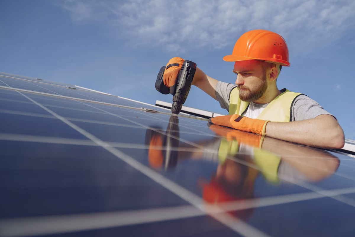 A worker in an orange hard hat, gloves and hi-vis vest uses a cordless drill on a solar panel.