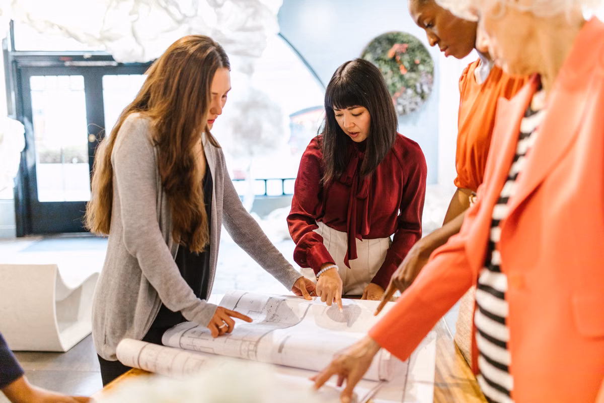 Four women gather around a table to look at and point to large printed architectural drawings.