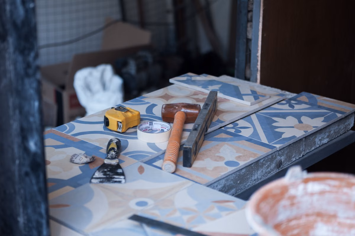Tiling tools, including a tape measure, rubber mallet, spirit level, masking tape and a scraper, laid on blue and cream patterned floor tiles on a workbench.