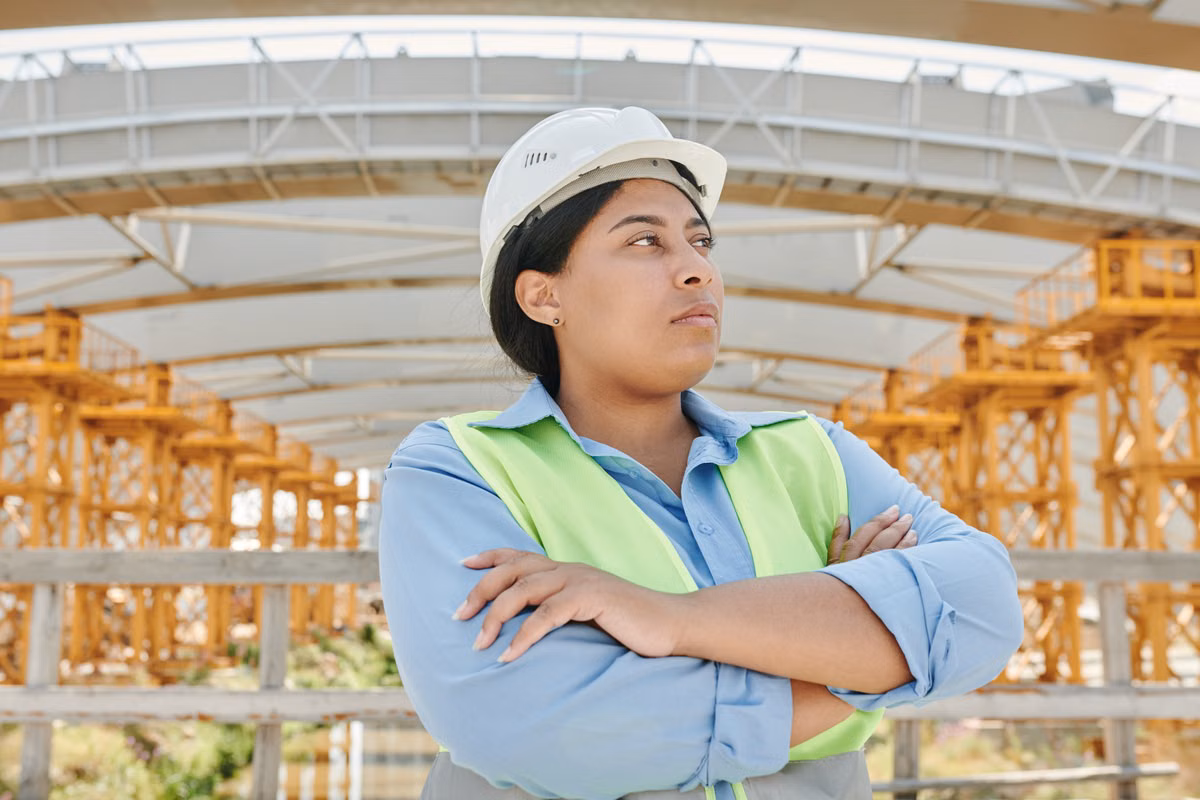 A woman wearing a white hard hat and a high-visibility vest stands with folded arms beneath a curved steel structure with yellow temporary support towers.