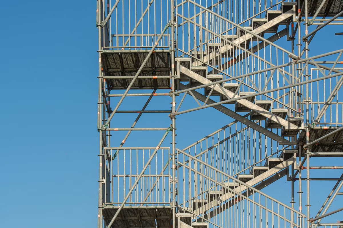 A galvanised steel scaffold stair tower with guardrails and mesh landings against a clear blue sky.