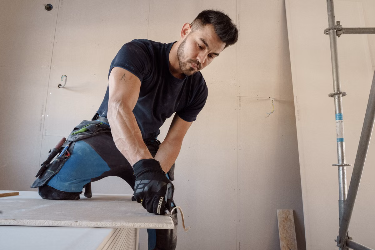 A worker in gloves and a tool belt kneels to cut a sheet of plasterboard inside a room lined with plasterboard, with a scaffold tower beside him.