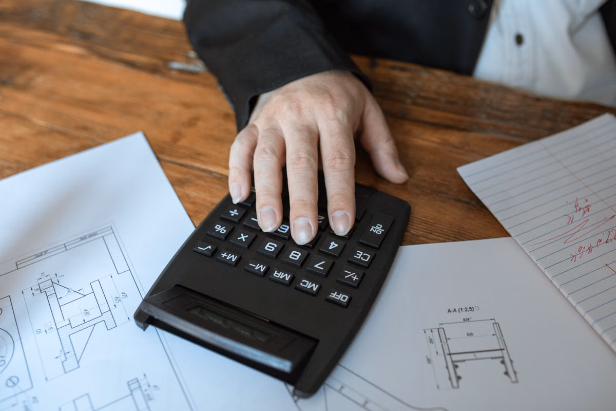 A hand uses a black calculator on a wooden desk next to technical drawings and a handwritten note.
