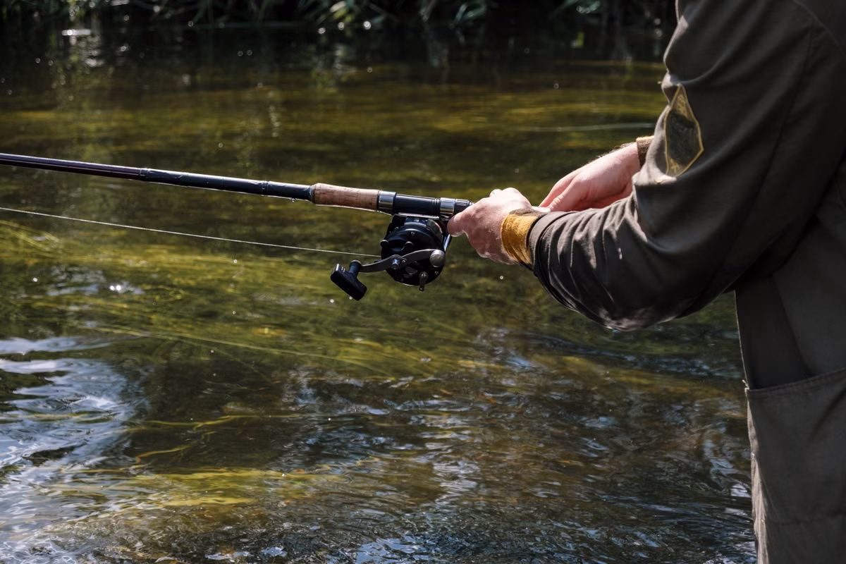 Close-up of an angler's hands holding a fishing rod and reel over a clear, shallow river.