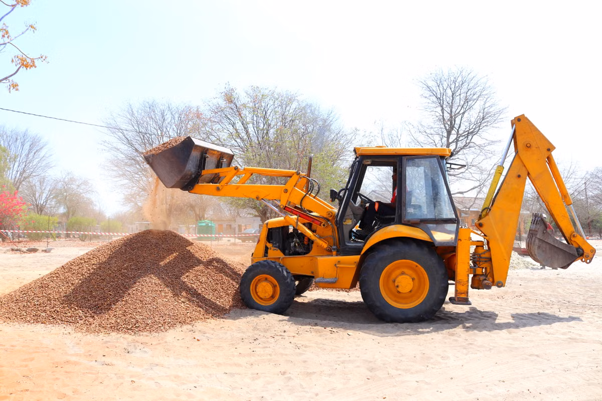 A yellow backhoe loader with an operator in the cab tips a bucket of gravel onto a pile on a sandy site.