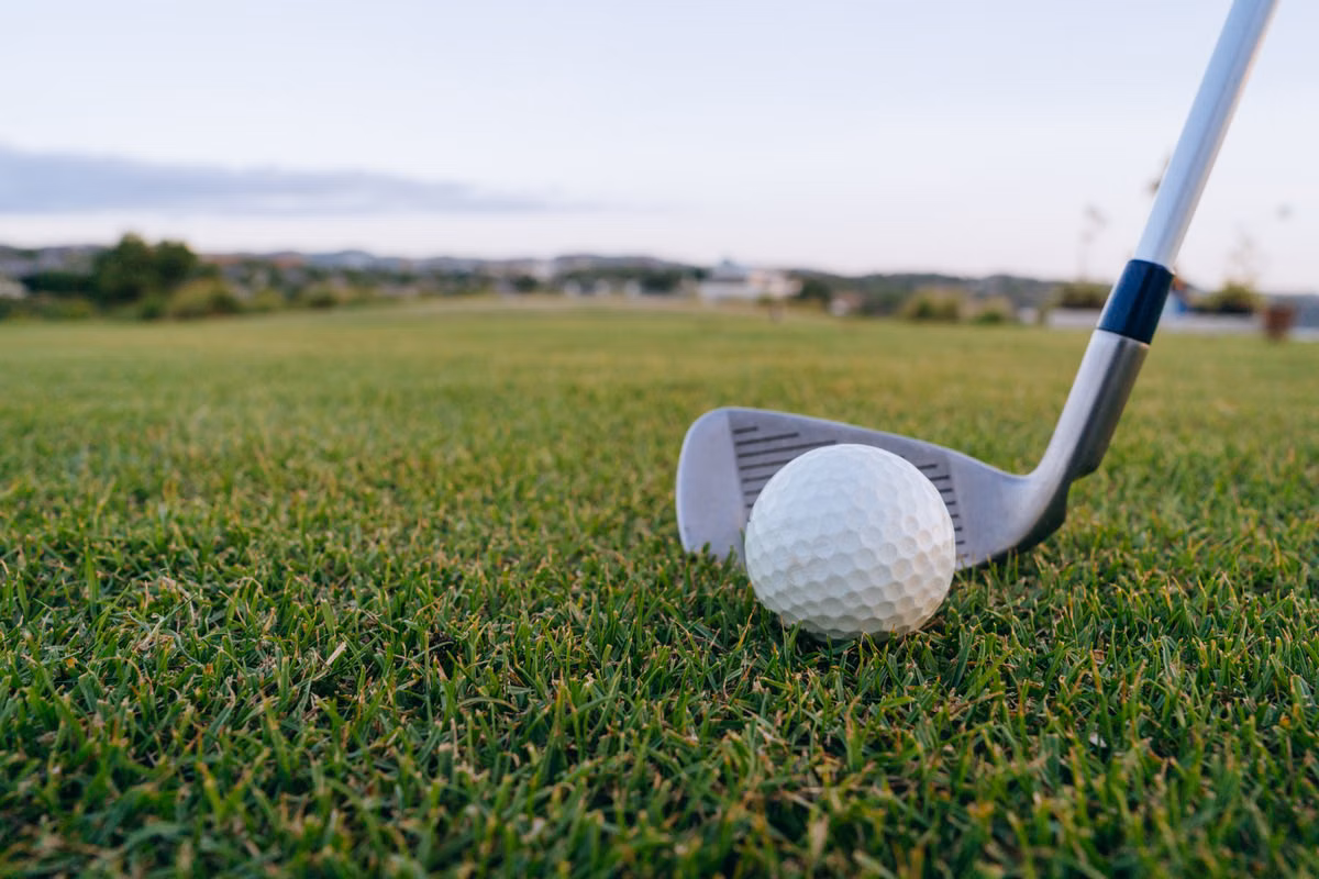 Close-up of a golf iron lined up behind a white golf ball on a grass fairway, with trees and buildings blurred in the distance.