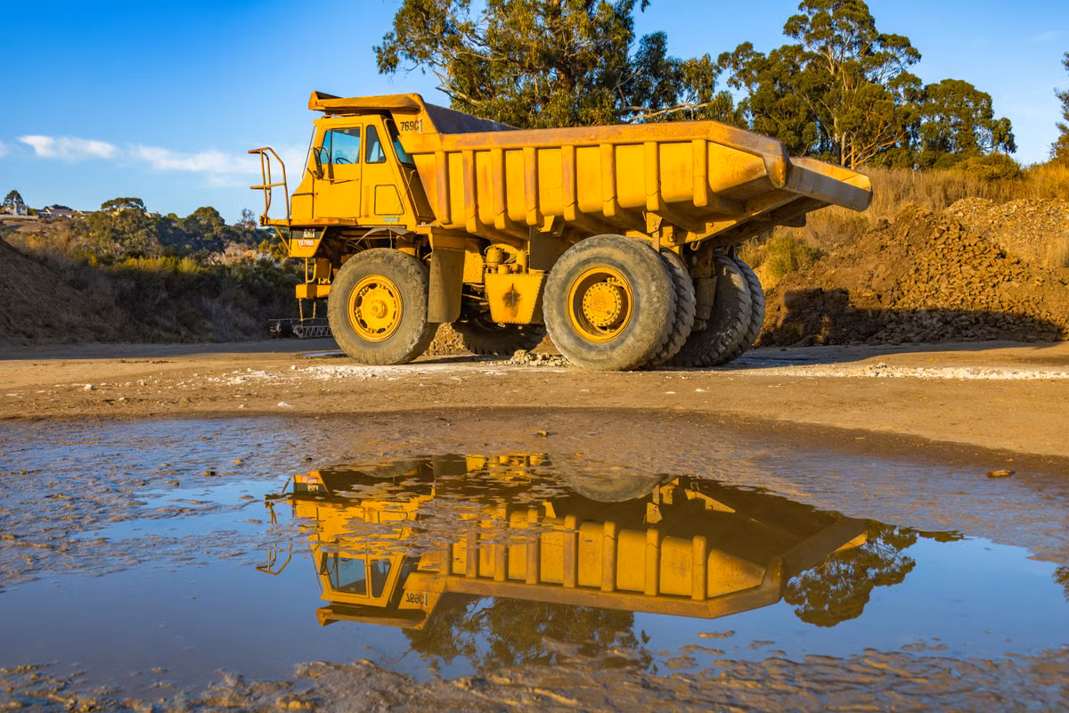 A yellow rigid dump truck parked on a quarry site, reflected in a puddle in the foreground.