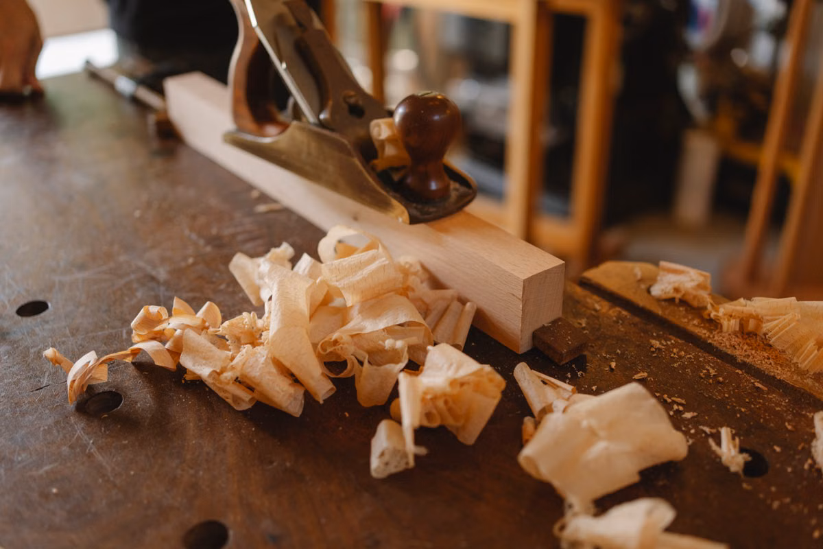A hand plane rests on a length of pale timber on a wooden workbench, surrounded by curled wood shavings.
